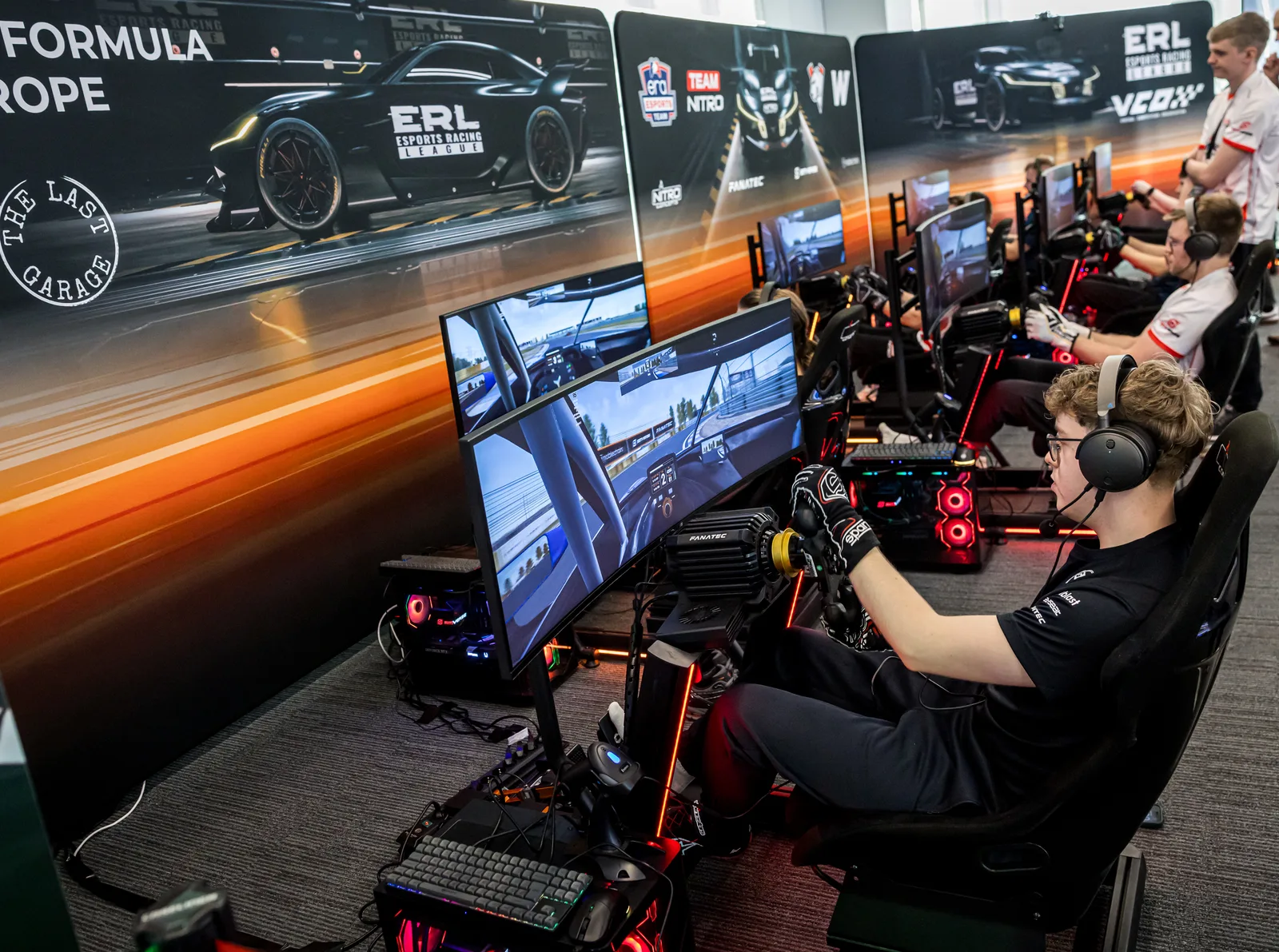 Drivers racing side by side on Fanatec sim rigs in the Esports Racing League area, with The Last Garage on the wall behind them